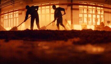 Two backlit distillery workers shoveling sorghum.